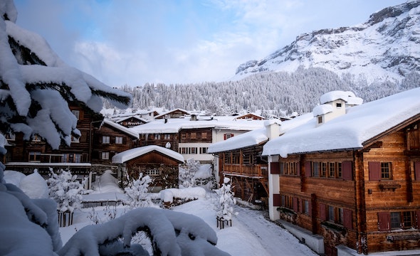 Snow covers the area and wooden cottages with snowy trees and a mountain side in the background