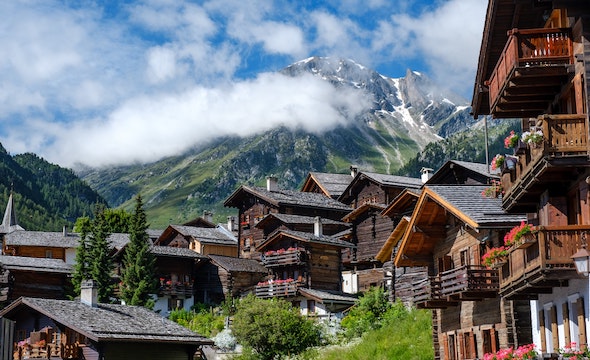 Wooden cottages dot a mountainside with a large mountain peak poking out of clouds is shown in the background