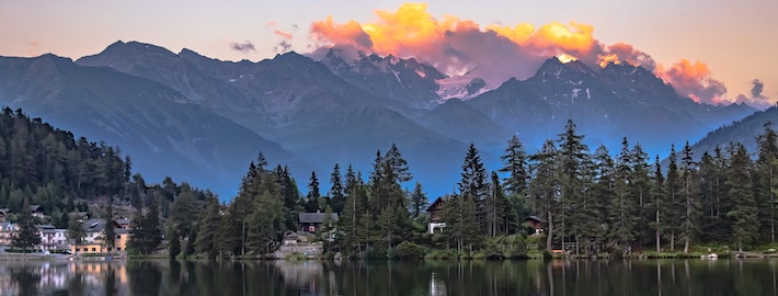 A scenic view of a sunset at a lake with large pine trees on the shore and an assortment of small cottages and buildings with large snowy mountains in the background